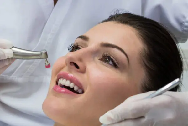 Patient smiling during a routine dental cleaning and exam while a dental professional uses a polishing handpiece and dental mirror to clean and inspect the teeth.